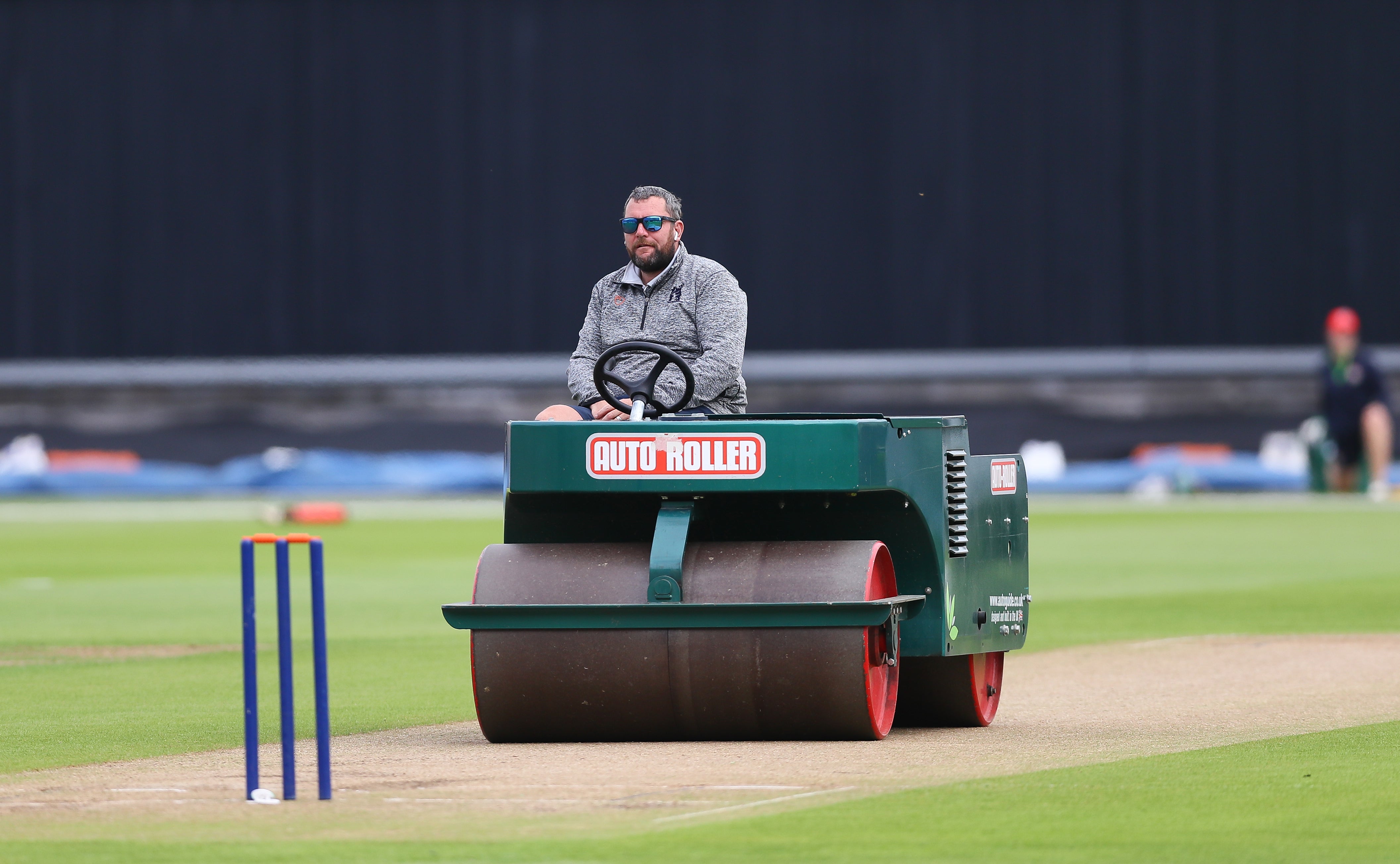 A groundskeeper driving a heavy motorized roller over a turf cricket pitch to flatten the surface
